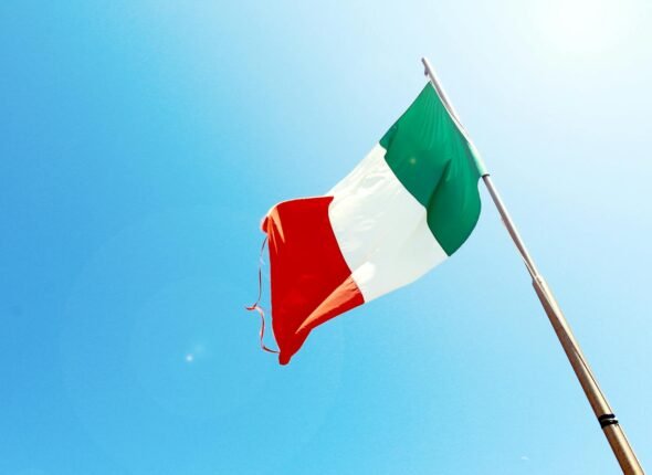 Vivid image of the Italian flag waving on a flagpole against a bright blue sky.